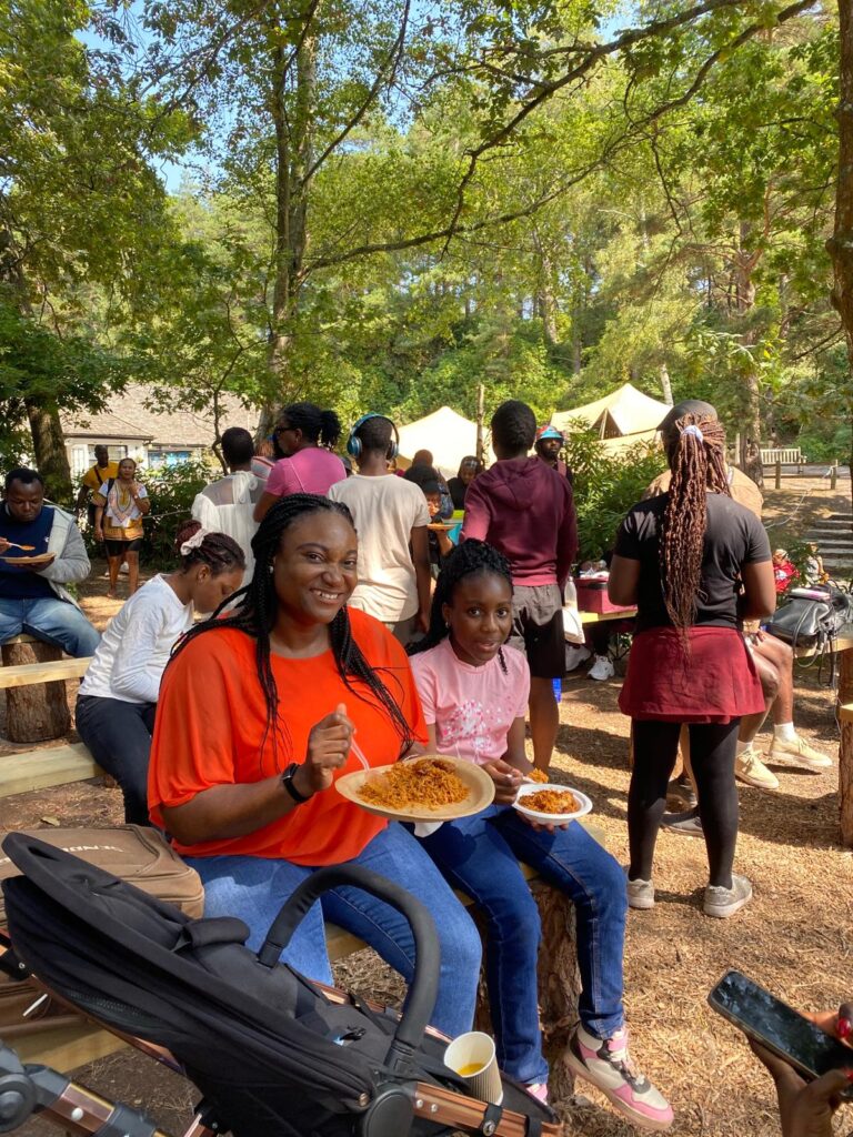 Two people sit outdoors with plates of food in a busy woodland picnic area, with others gathered in the background.