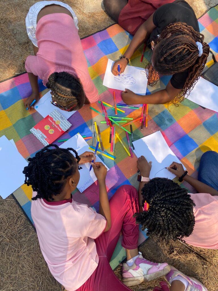 Children gather on a picnic blanket outdoors, drawing on large sheets of paper.