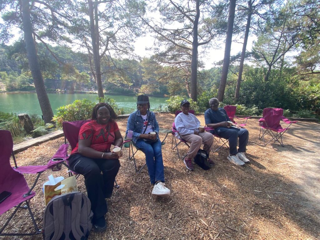 Four people sit on camping chairs beside a woodland lake, with tall trees and water visible in the background.