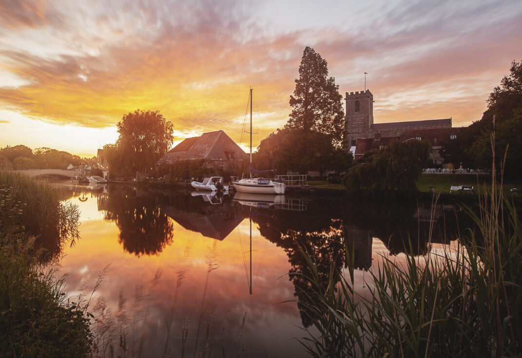 River with bright orange and pink sunset reflecting off the water with church and boats also reflected