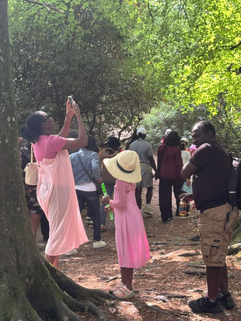 People gather on a shaded woodland path; one person takes a photo while a child stands nearby.