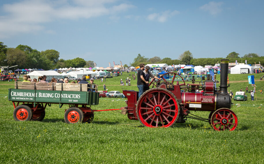 Vintage steam traction engine pulling a wagon across a showground, with visitors and event tents in the background at the Dorset Spring Show.