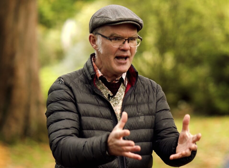 Man in green jacket talking animatedly, with blurred countryside background