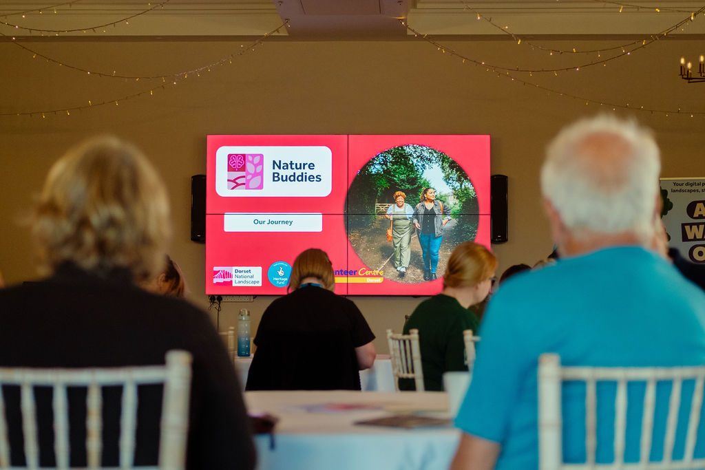 A screen showing a presentation with people sat on tables watching.
