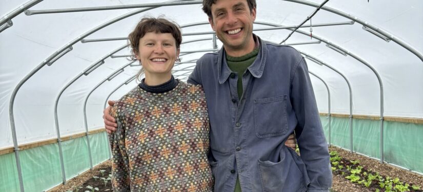 A farming couple smile inside their newly installed pollytunnel, standing in front of rows of vegetables