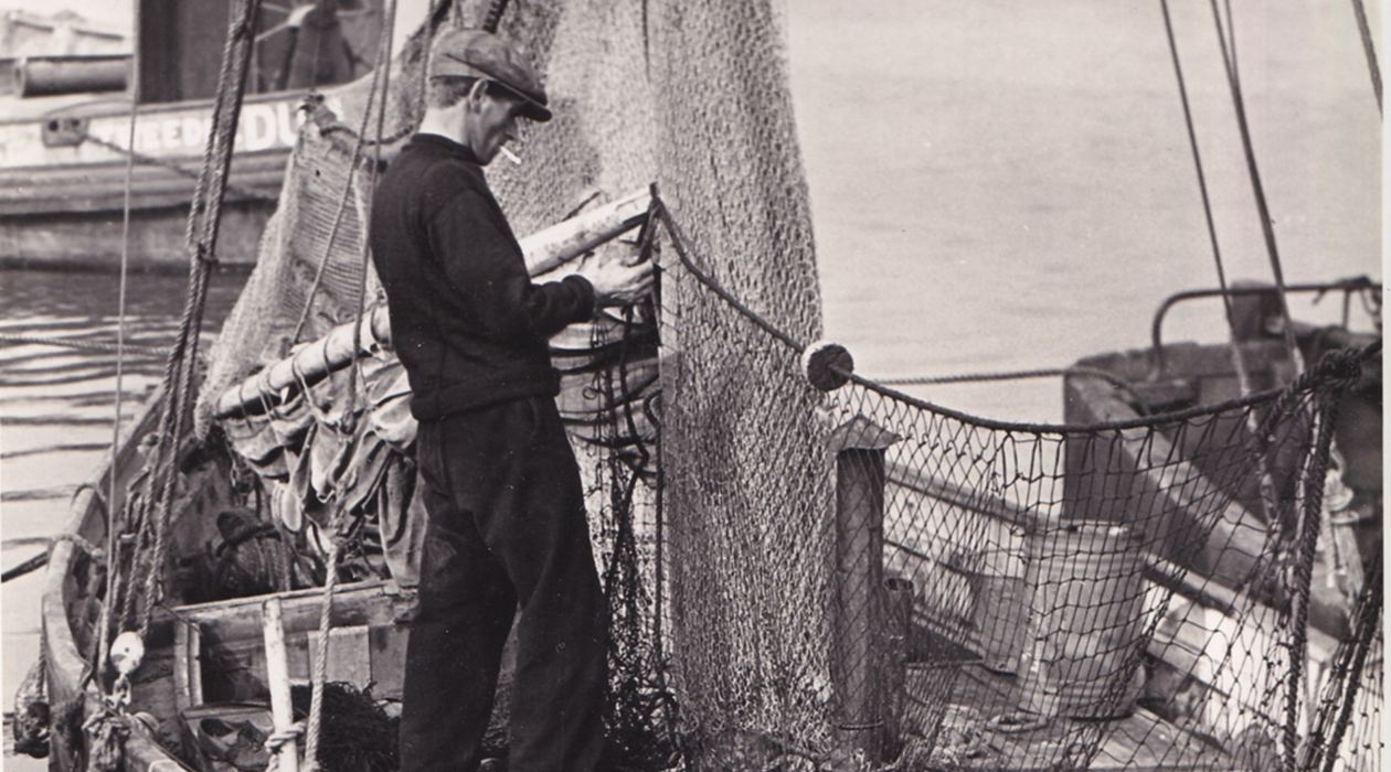 Historic black-and-white photograph showing a fisherman standing on a working fishing boat, repairing or handling large fishing nets. The harbour setting includes ropes, rigging and neighbouring vessels, illustrating traditional fishing life in Poole Harbour.