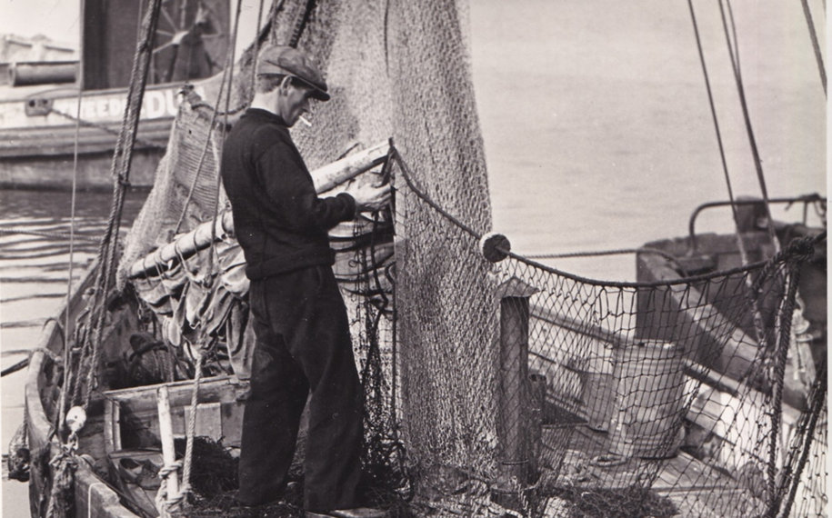 Historic black-and-white photograph showing a fisherman standing on a working fishing boat, repairing or handling large fishing nets. The harbour setting includes ropes, rigging and neighbouring vessels, illustrating traditional fishing life in Poole Harbour.