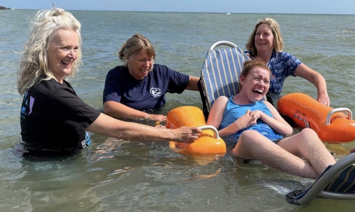 Participant using a floating beach-access wheelchair enjoys a supported sea swim in West Bay, assisted by three volunteers in shallow water.