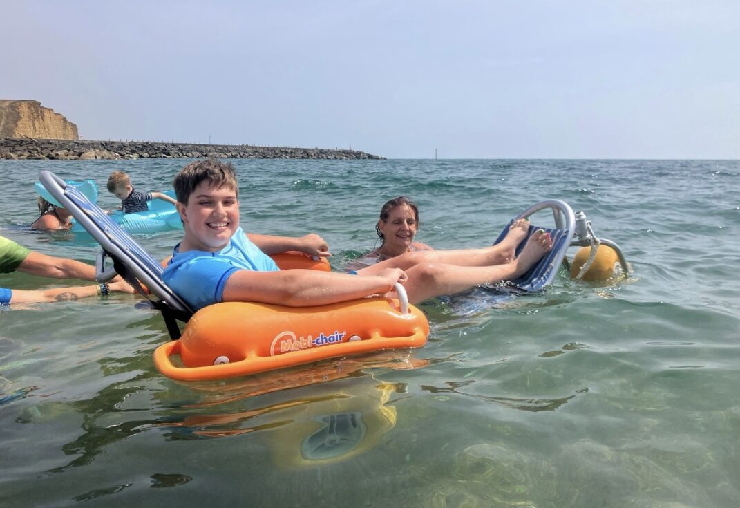 Participant relaxing in a floating beach wheelchair during an accessible sea swim in West Bay, accompanied by volunteers in the water near the shoreline.