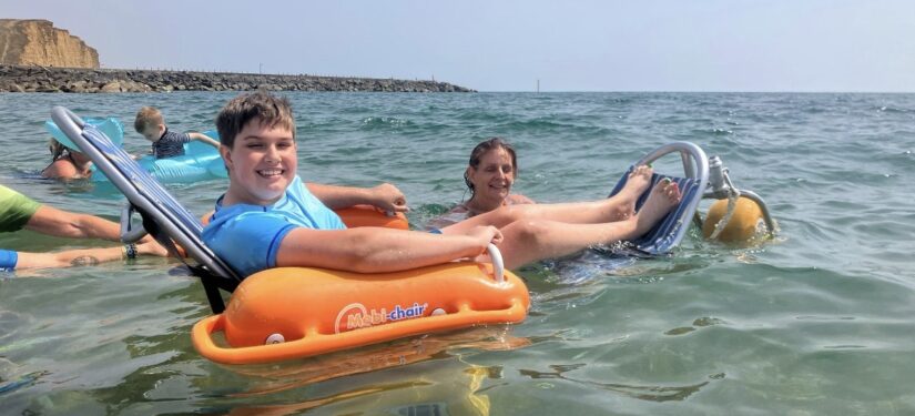 Participant relaxing in a floating beach wheelchair during an accessible sea swim in West Bay, accompanied by volunteers in the water near the shoreline.